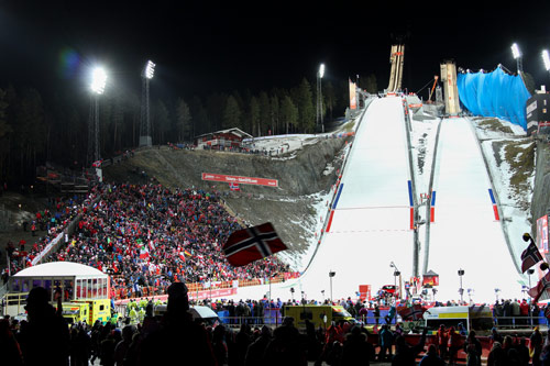 Falun ski jumps under floodlights at night, with a large crowd and a Norwegian flag in the foreground