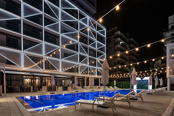 Hotel Nyota at night: facade with a lit triangular pattern, swimming pool and sun loungers