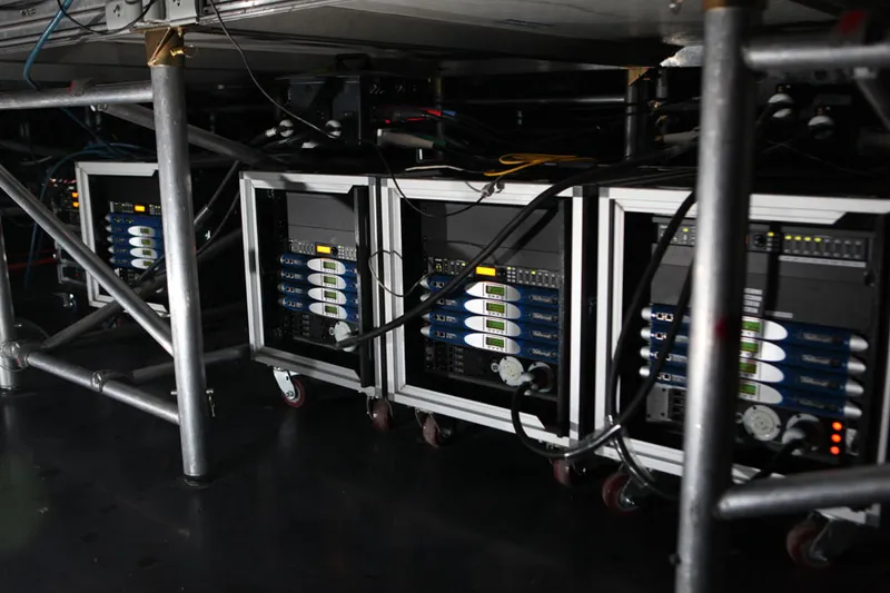 Rolling racks of Powersoft K Series amplifiers under the stage, among the scaffolding