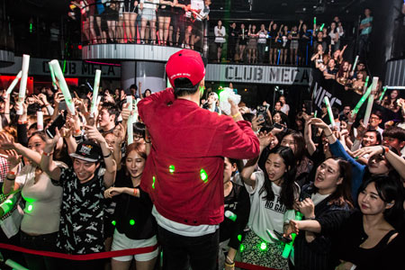 Performer in a red cap and jacket in front of a crowd holding glow sticks at Club Meca