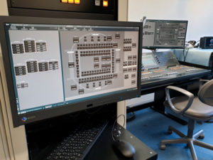 Control room monitor with device blocks laid out on a stadium plan, and a digital mixing console behind