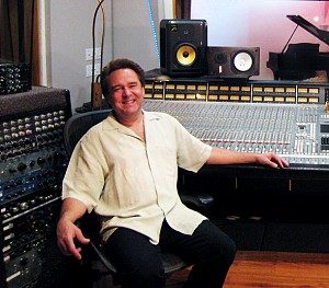 Man seated at a large analog recording console in a studio, with monitors on the meter bridge