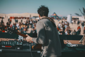 Guitarist on stage seen from behind, facing a daytime crowd among the desert dunes