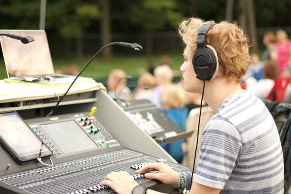 Young sound engineer with headphones at a digital mixing console outdoors, audience behind