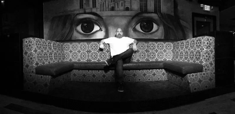 Man seated on a tiled booth below a large mural of a woman's eyes, black and white photo