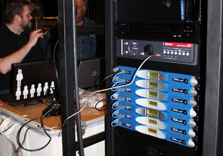 Rack with a Dolby cinema processor and Powersoft K Series amplifiers, technicians at laptops behind