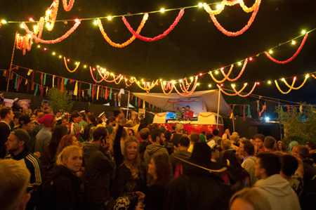 Crowd at night under string lights and garlands in front of the Hidden Disco DJ booth at Bestival