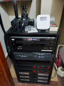 Equipment rack in a corner, with wireless microphone chargers on top and amplifiers at the bottom
