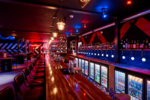 Long bar counter with stools, bottle shelves and drinks fridges under red and blue lighting