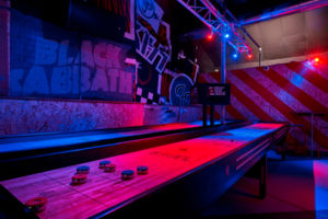 Shuffleboard tables under red and blue lighting, with a Black Sabbath mural on the wall