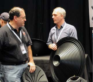Two men with event lanyards talking next to large black loudspeaker cones