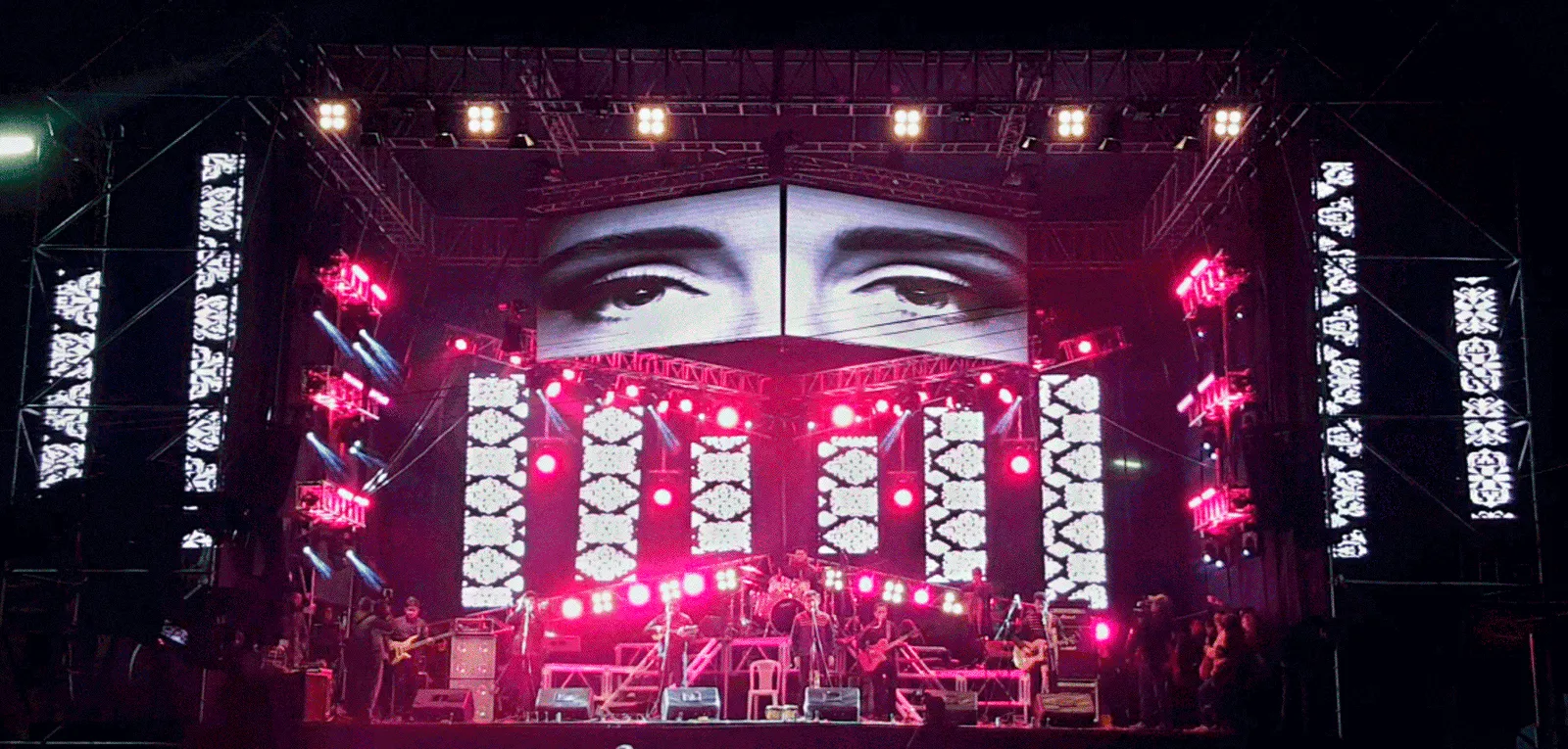 Concert stage at night with LED screens showing a pair of eyes, pink lights and a band performing