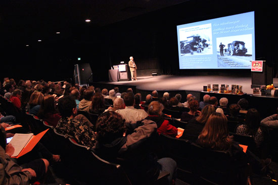 Full audience in the museum theater watching a presenter beside a screen with historic car photos