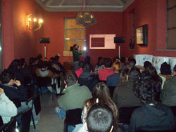 Seminar audience in a red-walled room with a presenter, loudspeakers on stands and chandeliers