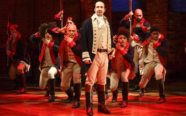 Hamilton cast on stage in 18th-century military costumes under red lighting