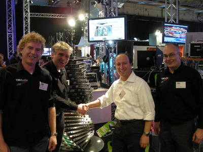 Handshake between TM Audio and Powersoft representatives at a trade show stand, beside a tower of stacked amplifiers