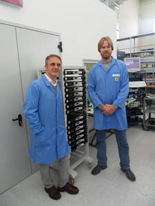 Two men in blue lab coats in an electronics production area, beside a trolley loaded with black units