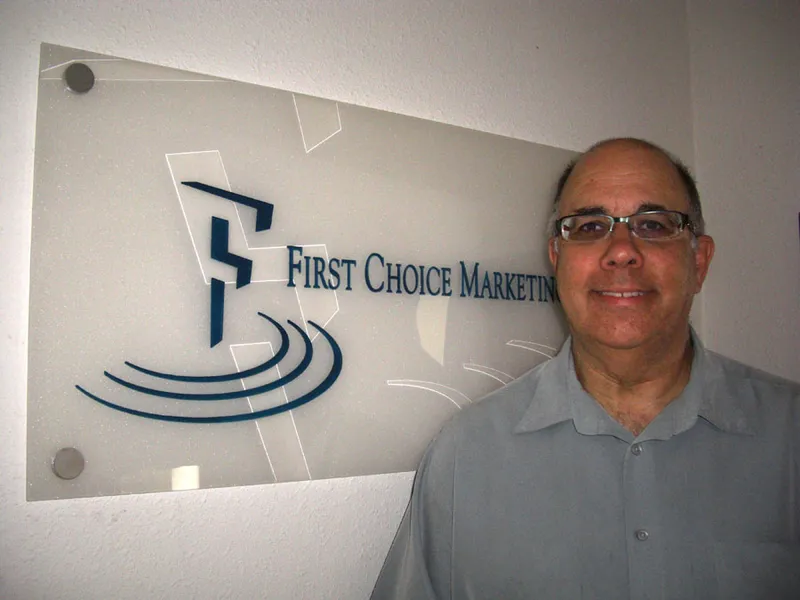 Man standing next to the First Choice Marketing logo sign on an office wall