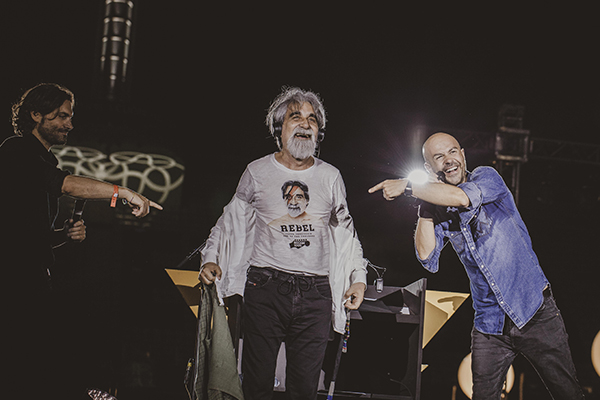 On stage at night, two men point at a white-bearded man wearing headphones and a Rebel t-shirt