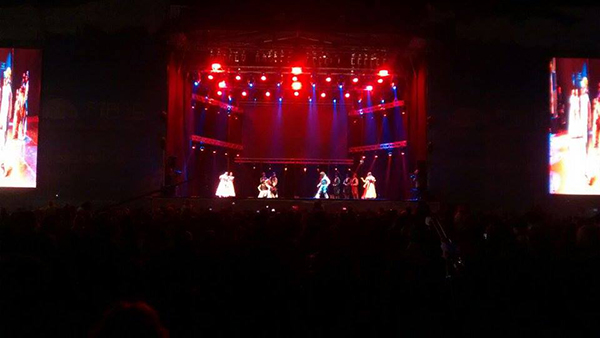 Performers in costume on the festival stage under red lights, with video screens on both sides