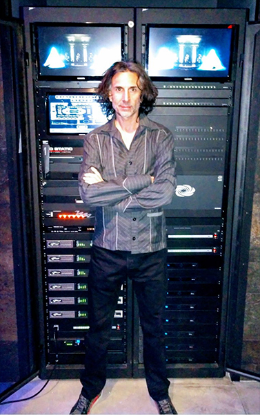 Man with arms crossed in front of an AV rack with screens and amplifiers